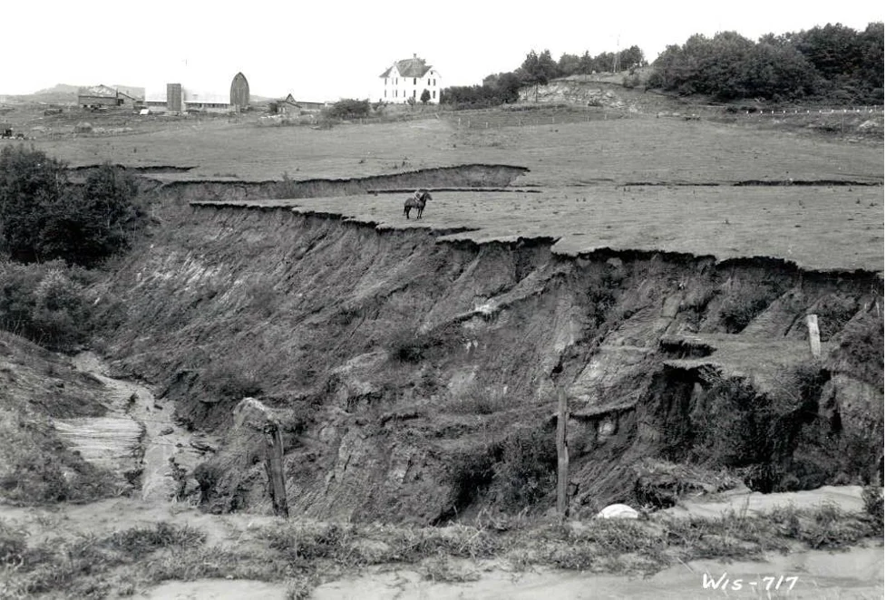 Black-and-white landscape with eroded ground, a distant horse and rider, and farm buildings